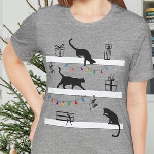 以下が含まれることがあります： 白棚に黒猫が描かれたデザインのグレーTシャツ。プレゼントとカラフルなストリングライトで飾られています。デザインは、お祝いのホリデーテーマです。シャツは柔らかい素材で作られています。