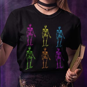 May include: Black t-shirt featuring six colorful skeleton illustrations. The skeletons are depicted in various colors, including purple, yellow, blue, green, orange, and pink. The design is a graphic print, perfect for Halloween or gothic-themed apparel.