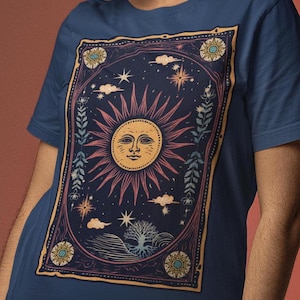 May include: A navy blue t-shirt with a vintage-style graphic of a sun with a face surrounded by stars, clouds, and floral elements. The graphic is framed in a gold border.