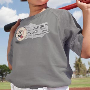 May include: A gray t-shirt with a baseball graphic and the word "Dinger" defined as a baseball home run. The shirt is being worn by a child holding a baseball bat.