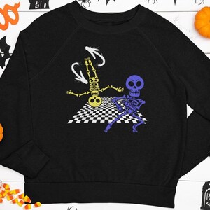 Op de afbeelding: Zwart sweatshirt met een afbeelding van twee dansende skeletten op een dambordvloer. Eén skelet is geel, de andere is paars. Het ontwerp heeft witte pijlaccenten, perfect voor Halloween.