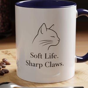 May include: White ceramic mug with a navy blue interior and handle. The mug features a black line drawing of a cat's profile and the text "Soft Life. Sharp Claws."