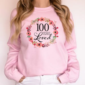 Peut inclure: Sweat-shirt rose avec un motif de couronne florale et le texte "100 Years Loved".