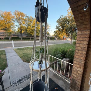 May include: A macrame plant hanger with three levels. The top level holds a black pot with a plant, the middle level has a wooden tray, and the bottom level has a gray pot. The hanger is suspended from a rope.