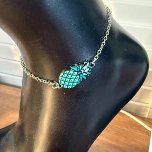 May include: Silver chain anklet with a turquoise and teal pineapple charm. The charm has a detailed design, and the anklet is displayed on a black mannequin foot. The anklet is a delicate piece of jewellery.
