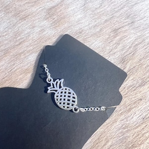 May include: A silver pineapple charm bracelet with a delicate chain. The charm is a cutout design of a pineapple with a detailed stem and leaves.