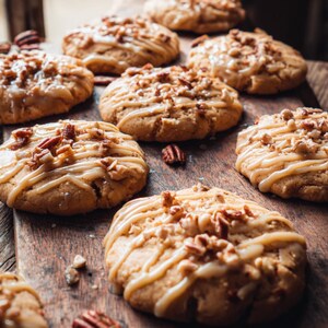 Könnte beinhalten: Auf einem Holzbrett werden mehrere Ahorn-Pekannuss-Kekse präsentiert. Jeder Keks hat eine hellbraune Basis, ist mit einer cremigen Glasur beträufelt und mit gehackten Pekannüssen belegt. Der Text "THE BEST MAPLE PECAN COOKIES" befindet sich in einem orangefarbenen Rechteck.