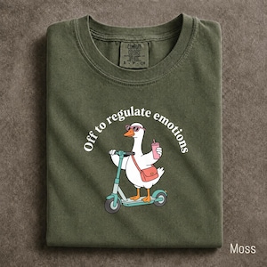 May include: A moss green t-shirt featuring a graphic of a white goose wearing pink sunglasses, riding a teal scooter, and carrying a pink shoulder bag while holding a pink drink. The text "Off to regulate emotions" is printed in white above the goose.