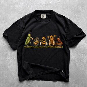 May include: A black short-sleeved t-shirt featuring a graphic print of five insects playing musical instruments. The illustration depicts a green grasshopper, a beetle with a guitar, a bee at a microphone, a cicada with maracas, and an ant on a xylophone.