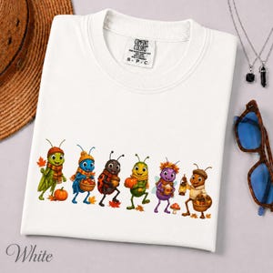 Peut inclure: Un t-shirt blanc avec une impression graphique de six insectes de dessin animé vêtus de vêtements d'automne. Les insectes tiennent des objets saisonniers comme des citrouilles, des glands et une lanterne, entourés de feuilles mortes.