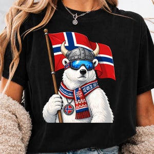 May include: A black t-shirt featuring a graphic of a white polar bear wearing a grey horned Viking helmet, blue ski goggles with a Norwegian flag pattern, and a red, white, and blue patterned scarf. The bear holds a Norwegian flag on a pole.