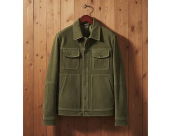 May include: Olive green jacket with four front pockets and a button-up closure. The jacket is hanging on a wooden hanger against a wooden background. White stitching outlines the pockets and seams.