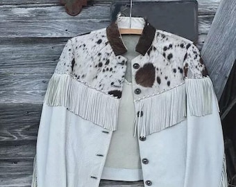 Könnte beinhalten: Eine weiße Lederjacke mit Kuhfell-Akzenten und Fransen. Die Jacke hat einen braunen Kragen und Manschetten und ist zugeknöpft. Die Jacke hängt an einem Holzbügel vor einem rustikalen Holzhintergrund.