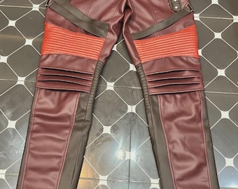 Peut inclure: Pantalon en similicuir bordeaux avec des accents rouges et marron. Le pantalon est doté d'une ceinture à boucle, de sangles décoratives et de détails côtelés sur les genoux. Une petite étiquette indique "LEATHER".