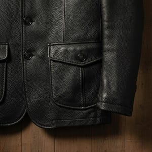 May include: Close-up of a black leather jacket with a textured finish. The jacket features two front pockets with button closures, a notched lapel, and buttoned cuffs. The jacket is a classic style.