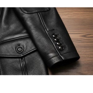May include: Close-up of a black leather jacket. The jacket features a textured surface, a buttoned pocket, and a sleeve with four black buttons. The jacket is on a wooden surface.