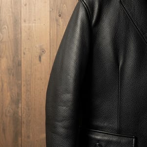 May include: Close-up of a black leather jacket with a textured surface. The jacket has a classic design with a notched lapel and a single button closure. The leather appears to be high-quality and durable.