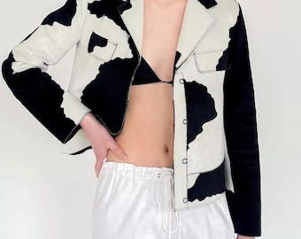 May include: A black and white cow print cropped jacket with a button closure and a single pocket on the left side. The jacket is worn over a black bra and white pants.