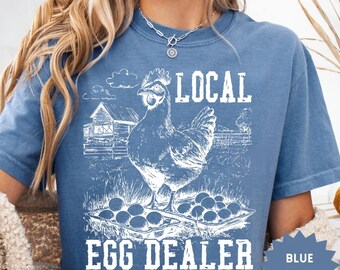 May include: Blue jean colored t-shirt with a white graphic of a hen, eggs, and a barn. The text on the shirt reads "LOCAL EGG DEALER". A blue starburst tag reads "BLUE JEAN".