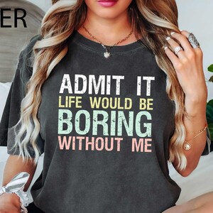 Op de afbeelding: Donkergrijs t-shirt met de tekst "ADMIT IT LIFE WOULD BE BORING WITHOUT ME" in wit, geel, groen en roze. Het shirt heeft een casual stijl.