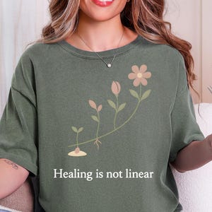 May include: A moss green short-sleeve t-shirt featuring a graphic of a plant growing in stages from a seed to a flower. Below the plant illustration, the text reads, "Healing is not linear" in a clean, white, sans-serif font.