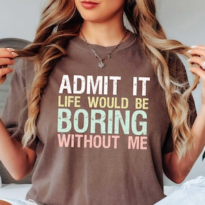 Op de afbeelding: Een bruin T-shirt met de tekst "ADMIT IT LIFE WOULD BE BORING WITHOUT ME" in wit, geel, groen en roze. Het shirt wordt gedragen met lichtblauwe jeans. Het thema is vrijetijdskleding.