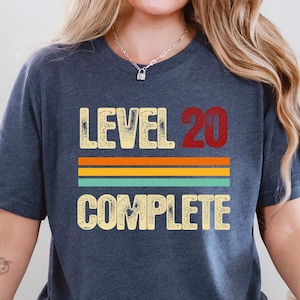 May include: A navy blue t-shirt with the text "LEVEL 20 COMPLETE" in a distressed, vintage style. The text is layered over horizontal stripes in orange, teal, and yellow. The shirt is a casual, unisex style.