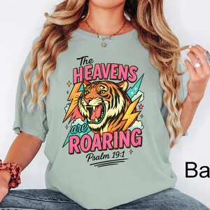 May include: A light green t-shirt with a colorful graphic of a tiger's face, lightning bolts, and the text "The Heavens are Roaring" in pink and white. The text "Psalm 19:1" is below the graphic.