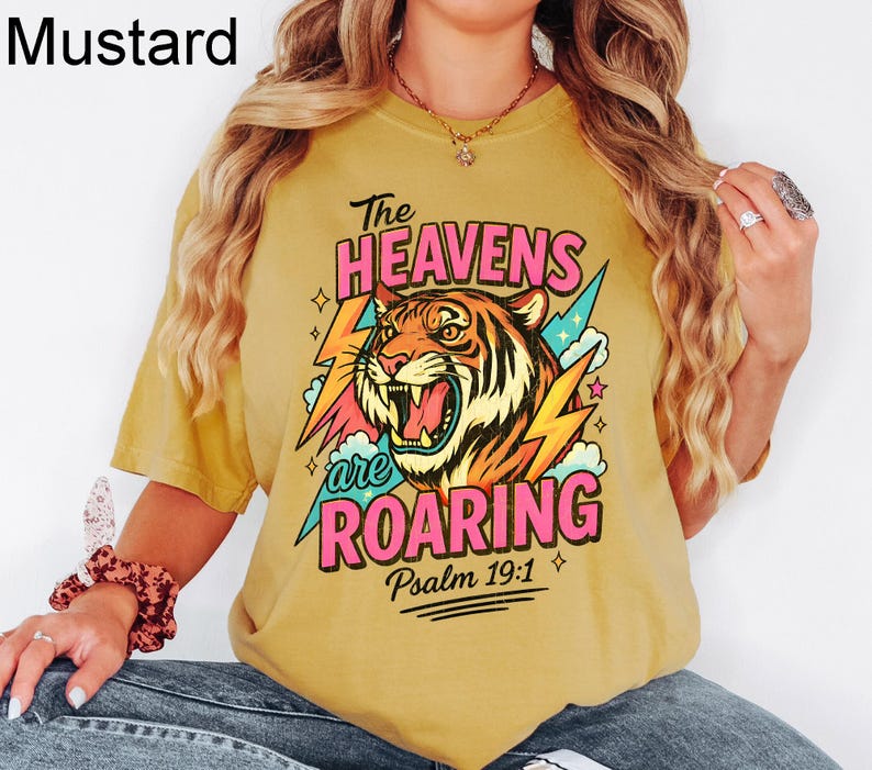 May include: Mustard-colored t-shirt with a graphic of a roaring tiger, lightning bolts, and the text "The Heavens are Roaring" in pink and green. The text "Psalm 19:1" is also included.