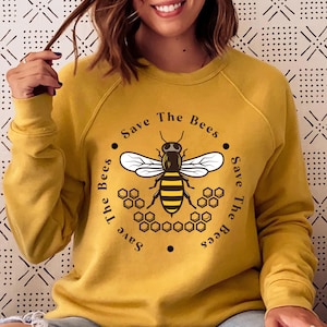 May include: Mustard yellow sweatshirt with a graphic of a bee and the text "Save The Bees" in a circular design. The bee is black and yellow with white wings, surrounded by honeycomb shapes. A casual, comfortable top.