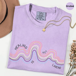 May include: An orchid-colored t-shirt with a crew neckline and a Comfort Colors label. The shirt features a graphic of wavy lines in pink, yellow, blue, and purple with the black text "HEALING IS NOT LINEAR" printed across the wavy design.