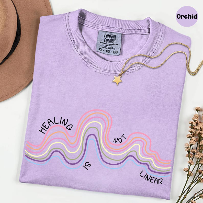 May include: An orchid-colored t-shirt with a crew neckline and a Comfort Colors label. The shirt features a graphic of wavy lines in pink, yellow, blue, and purple with the black text "HEALING IS NOT LINEAR" printed across the wavy design.