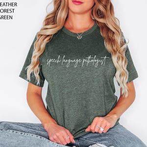 May include: Heather forest green t-shirt with the text "speech language pathologist" in a white script. The shirt is a crew neck style and has short sleeves. The shirt is made of a soft material.