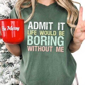 Op de afbeelding: Een rode keramische mok met de tekst "Be Merry" in witte letters wordt voor een groen t-shirt gehouden. Het t-shirt heeft de tekst "ADMIT IT LIFE WOULD BE BORING WITHOUT ME" in wit, geel, blauw en roze.