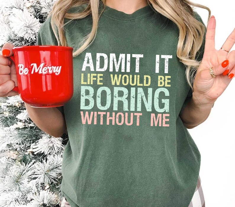 Op de afbeelding: Een rode keramische mok met de tekst "Be Merry" in witte letters wordt voor een groen t-shirt gehouden. Het t-shirt heeft de tekst "ADMIT IT LIFE WOULD BE BORING WITHOUT ME" in wit, geel, blauw en roze.