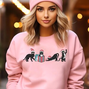 May include: Pink sweatshirt featuring a festive design of black cats wearing Santa hats. One cat is in a gift box, another is tangled in lights, and the others are in playful poses. The sweatshirt is a solid pink color.