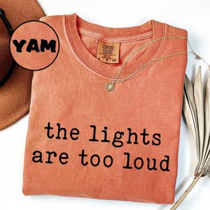 May include: Coral-colored t-shirt with the text "the lights are too loud" in black. A gold necklace with a pendant is draped on the shirt. A brown hat and dried plant are partially visible in the background. The word "YAM" is in a circle.