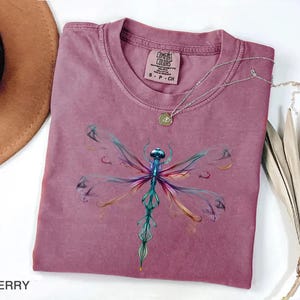 May include: A berry-colored short-sleeve t-shirt featuring a graphic of a dragonfly with iridescent, multi-colored wings in shades of blue, purple, and gold. The shirt has a crew neckline and a Comfort Colors brand label printed on the inner collar.