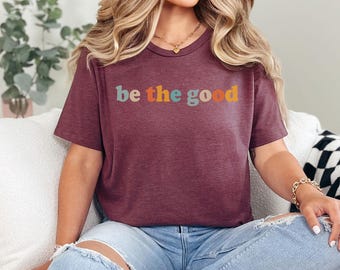 May include: A maroon t-shirt with the phrase "be the good" printed across the front in teal, orange, and yellow lettering. The shirt has a crew neck and short sleeves. The model is wearing blue jeans.
