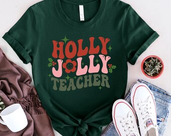 May include: Dark green t-shirt with the words "HOLLY JOLLY TEACHER" in red, pink, and green. The design includes a smiling flower and holly accents. White sneakers and a small potted plant are visible.