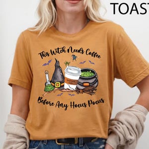 May include: Mustard-colored t-shirt with a Halloween-themed graphic. The design includes a witch's hat, a coffee cup, a cauldron, and the text "This Witch Needs Coffee Before Any Hocus Pocus."