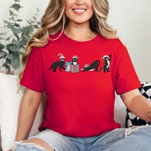 May include: A red t-shirt featuring a festive design with four black cats wearing Santa hats. The cats are engaged in various activities, including opening a gift and playing with Christmas lights. The shirt is made of a soft material.