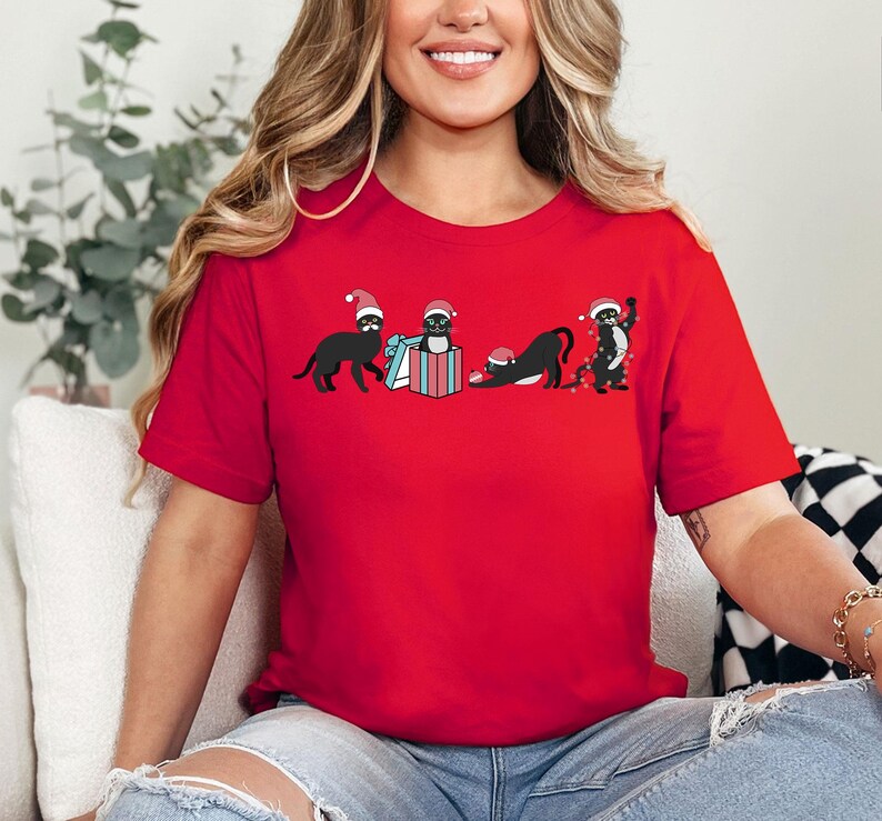 May include: A red t-shirt featuring a festive design with four black cats wearing Santa hats. The cats are engaged in various activities, including opening a gift and playing with Christmas lights. The shirt is made of a soft material.