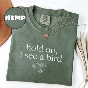 May include: Olive green t-shirt with the text "hold on, i see a bird" and a graphic of binoculars. A gold necklace with a pendant is on the shirt. The word "HEMP" is in a circle in the upper left corner.