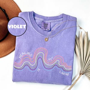 May include: A violet short-sleeve t-shirt with a crew neckline and a graphic print featuring wavy lines in pink, yellow, and light blue. The text "HEALING IS NOT LINEAR" is printed in white. A small circular inset label reads "VIOLET".