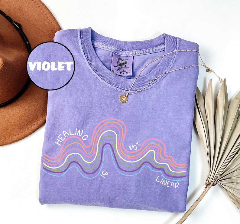 May include: A violet short-sleeve t-shirt with a crew neckline and a graphic print featuring wavy lines in pink, yellow, and light blue. The text "HEALING IS NOT LINEAR" is printed in white. A small circular inset label reads "VIOLET".