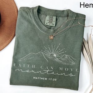 May include: Olive green t-shirt with a mountain and sun graphic, and the text "Faith Can Move Mountains" and "Matthew 17:20". A silver necklace with a pendant is draped across the shirt. The shirt is folded.