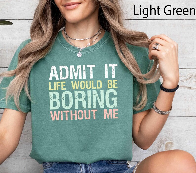 Op de afbeelding: Lichtgroen T-shirt met de tekst "GEEF HET TOE, HET LEVEN ZOU ZONDER MIJ VERVELEND ZIJN" in wit, geel, lichtblauw en roze. Het shirt is effen van kleur en heeft een ronde hals.