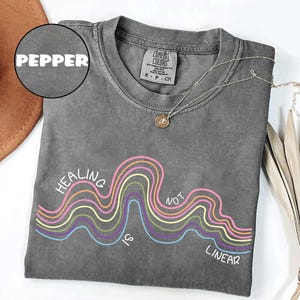 May include: A pepper-colored short-sleeve t-shirt featuring a graphic of multi-colored wavy lines and the text "HEALING IS NOT LINEAR" in white. The shirt has a crew neckline and a Comfort Colors brand label on the inside of the collar.