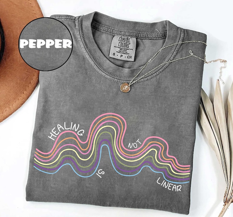 May include: A pepper-colored short-sleeve t-shirt featuring a graphic of multi-colored wavy lines and the text "HEALING IS NOT LINEAR" in white. The shirt has a crew neckline and a Comfort Colors brand label on the inside of the collar.